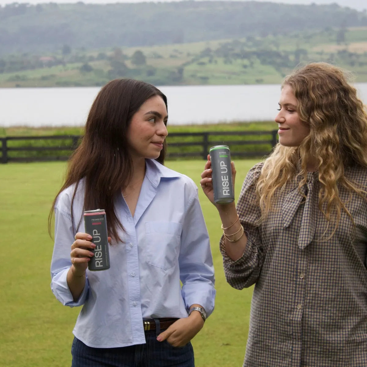Dos amigas conversando en el pasto junto a un lago, cada una con su lata de Rise Up Frambuesa y Citrus